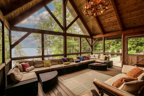 Modern wood cabin with illuminated windows and wraparound deck at dusk, surrounded by evergreen trees and landscaping.